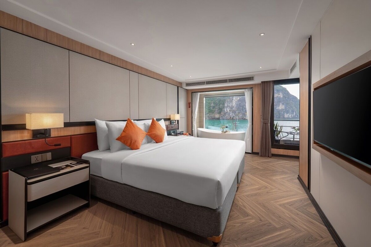 A hotel room with a large bed, a bedside table, a TV, a window with a view of the sea, and a balcony.