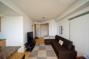 A modern living room with a brown sofa, a flat-screen TV, and a kitchen area with a granite countertop. - Fairview Downtown (Toronto)