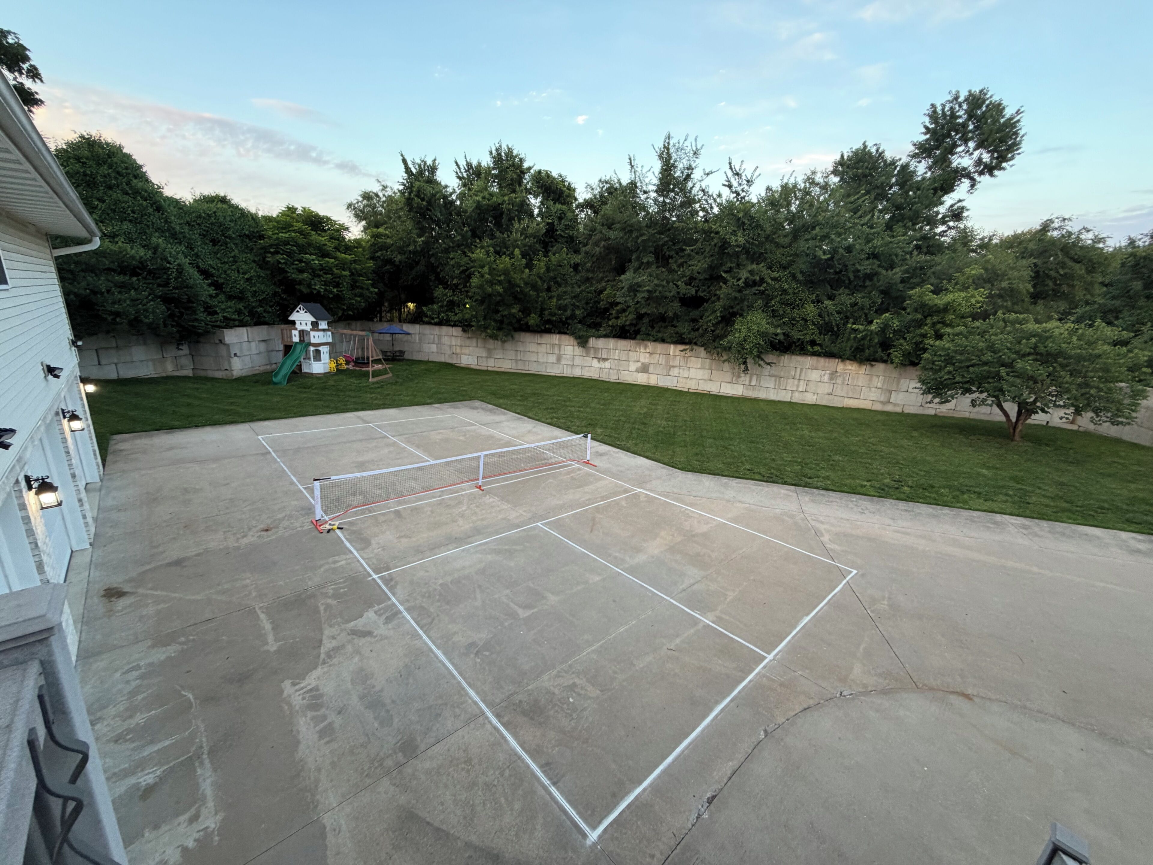 A backyard tennis court with a net, surrounded by a stone wall and trees.