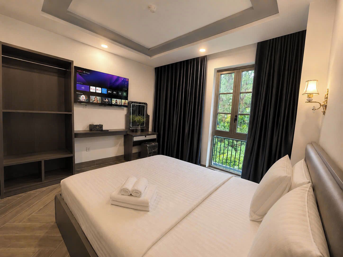 A hotel room with a bed, a TV mounted on the wall, a desk with a chair, and a window with curtains.