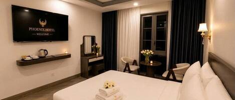 A hotel room with a bed, a TV mounted on the wall, a desk with a chair, a small table with a lamp, and a window with curtains.