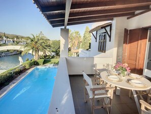 Zona de piscina amb taula preparada per dinar, terrassa coberta i vista a un cos d'aigua i vegetació.