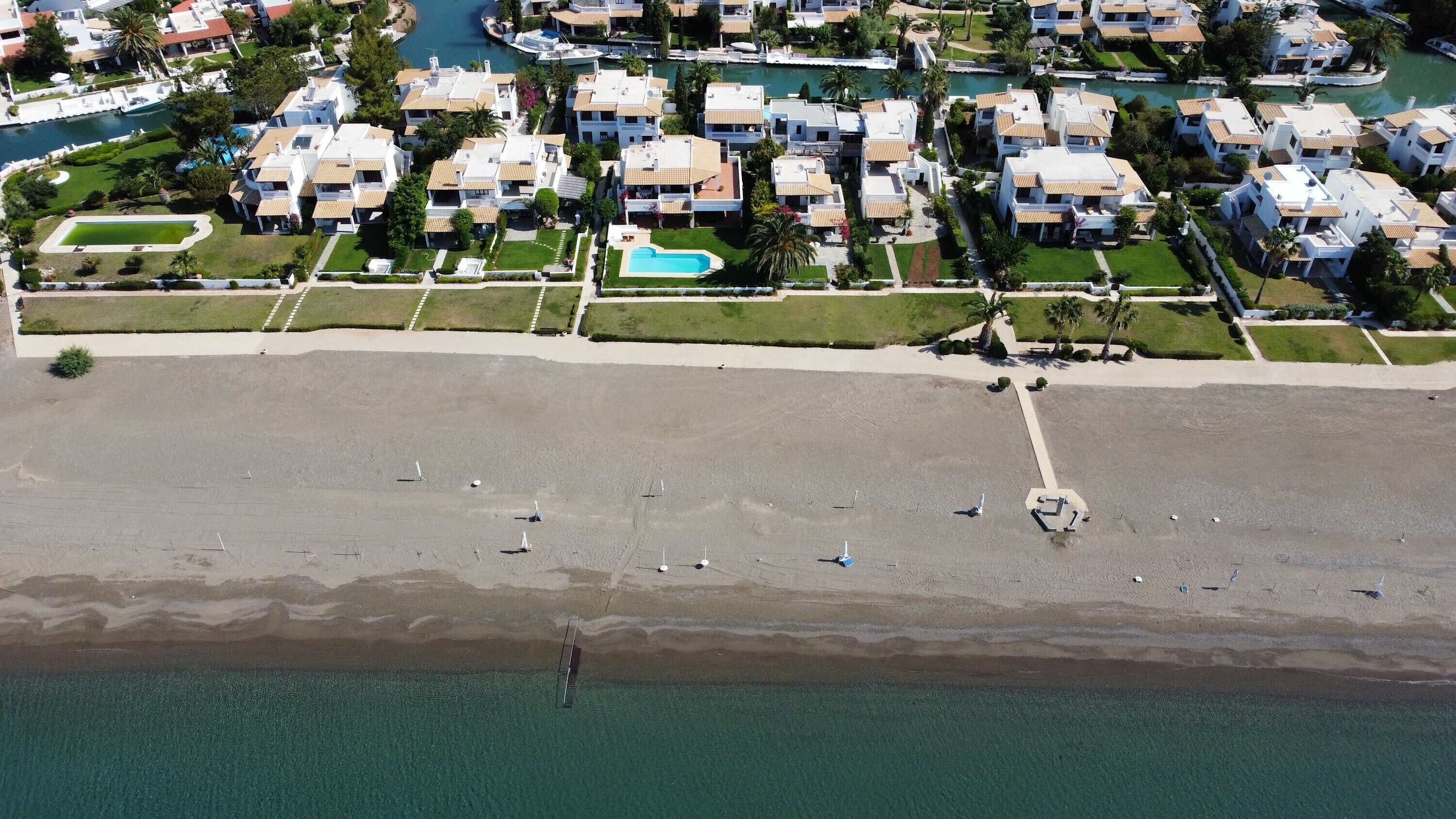 Vista aérea de uma área residencial costeira com vários edifícios brancos, uma praia de areia e um corpo de água azul e límpido.
