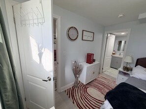A hotel room with a bed, a bedside table, a mirror, a clock, and a door with a coat rack. - Parkview - A Cozy Gem (Mississauga)