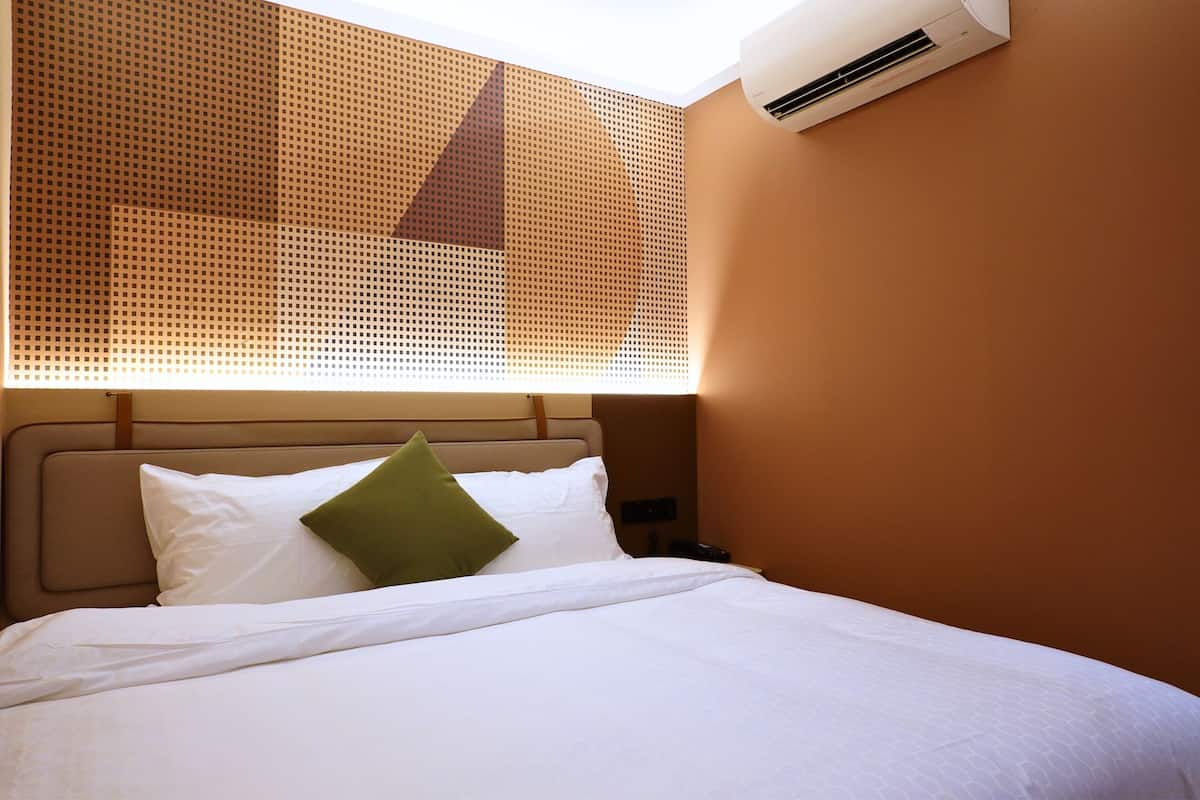 A hotel room with a bed, a headboard, a nightstand, and an air conditioning unit mounted on the wall.