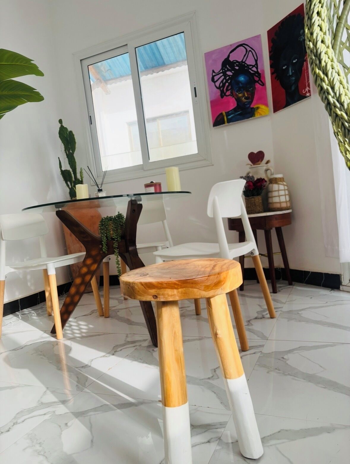 Une pièce avec une table à plateau en verre, des chaises blanches, un tabouret en bois, une plante succulente et des œuvres d’art accrochées au mur.