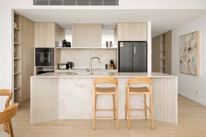 A modern kitchen with a central island, built-in appliances, and wooden bar stools. - Amaya Coastal Luxe | Pool & Panoramic Views (Gold Coast)