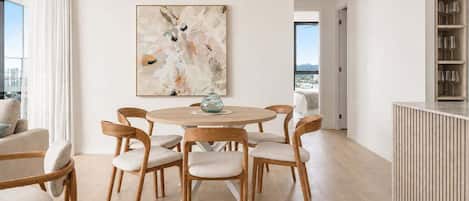 A modern dining area with a round table and six chairs, a large abstract painting on the wall, and a view of the city through the window.