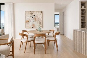 A modern dining area with a round table and six chairs, a large abstract painting on the wall, and a view of the city through the window. - Amaya Coastal Luxe | Pool & Panoramic Views (Gold Coast)