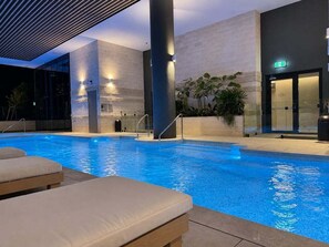 An indoor swimming pool with lounge chairs, a glass wall, and a modern architectural design. - Amaya Coastal Luxe | Pool & Panoramic Views (Gold Coast)