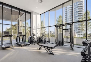A modern gym with treadmills, elliptical machines, and weight benches, large windows, and a view of high-rise buildings. - Amaya Coastal Luxe | Pool & Panoramic Views (Gold Coast)
