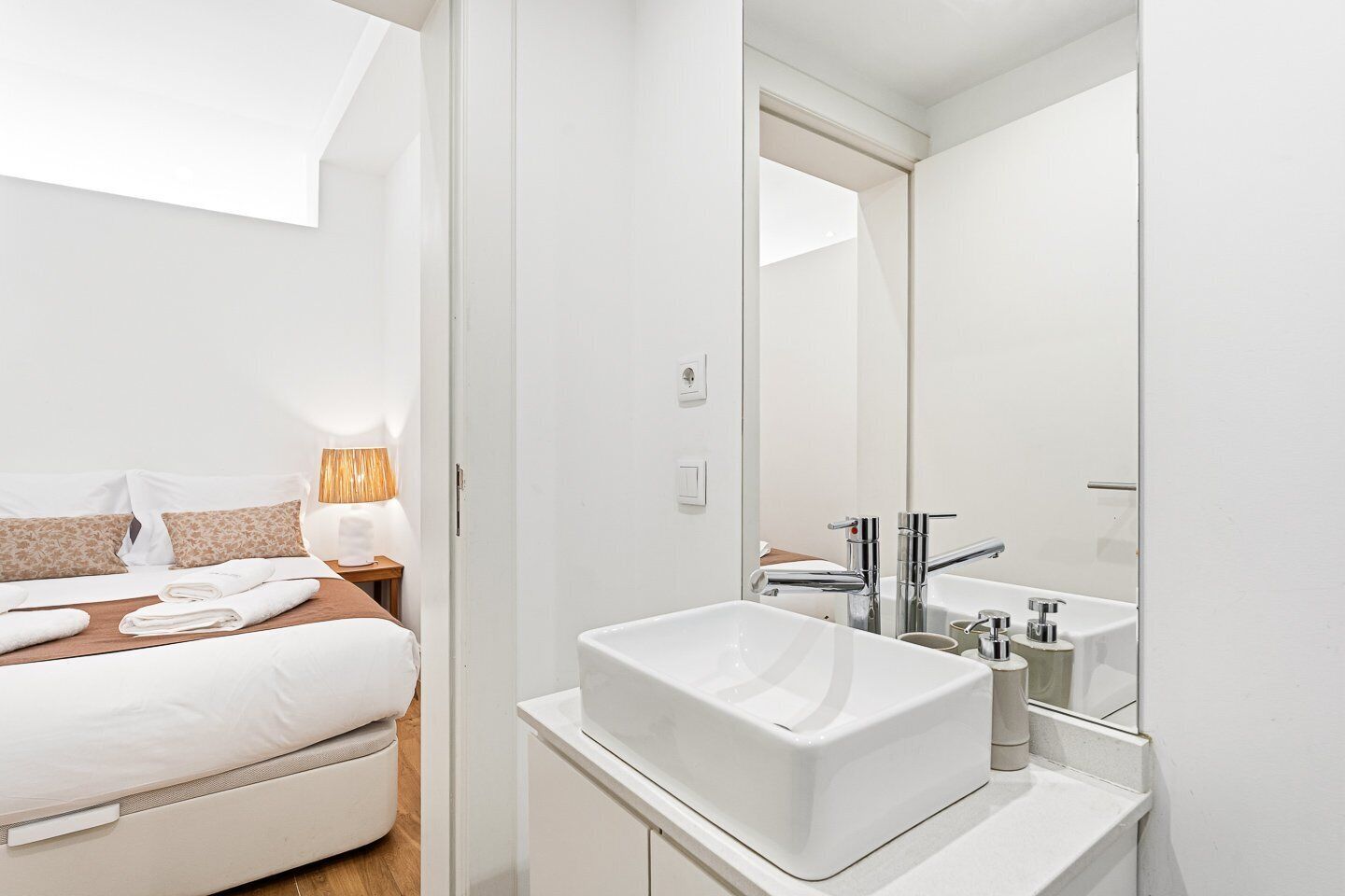 Una habitación de hotel moderna con cama, lámpara de noche y un espejo grande sobre un lavamanos blanco.