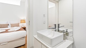 Habitació d'hotel moderna amb llit, tauleta de nit i un gran mirall sobre un lavabo blanc.