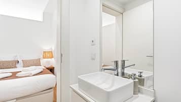 Una habitación de hotel moderna con cama, lámpara de noche y un espejo grande sobre un lavamanos blanco.