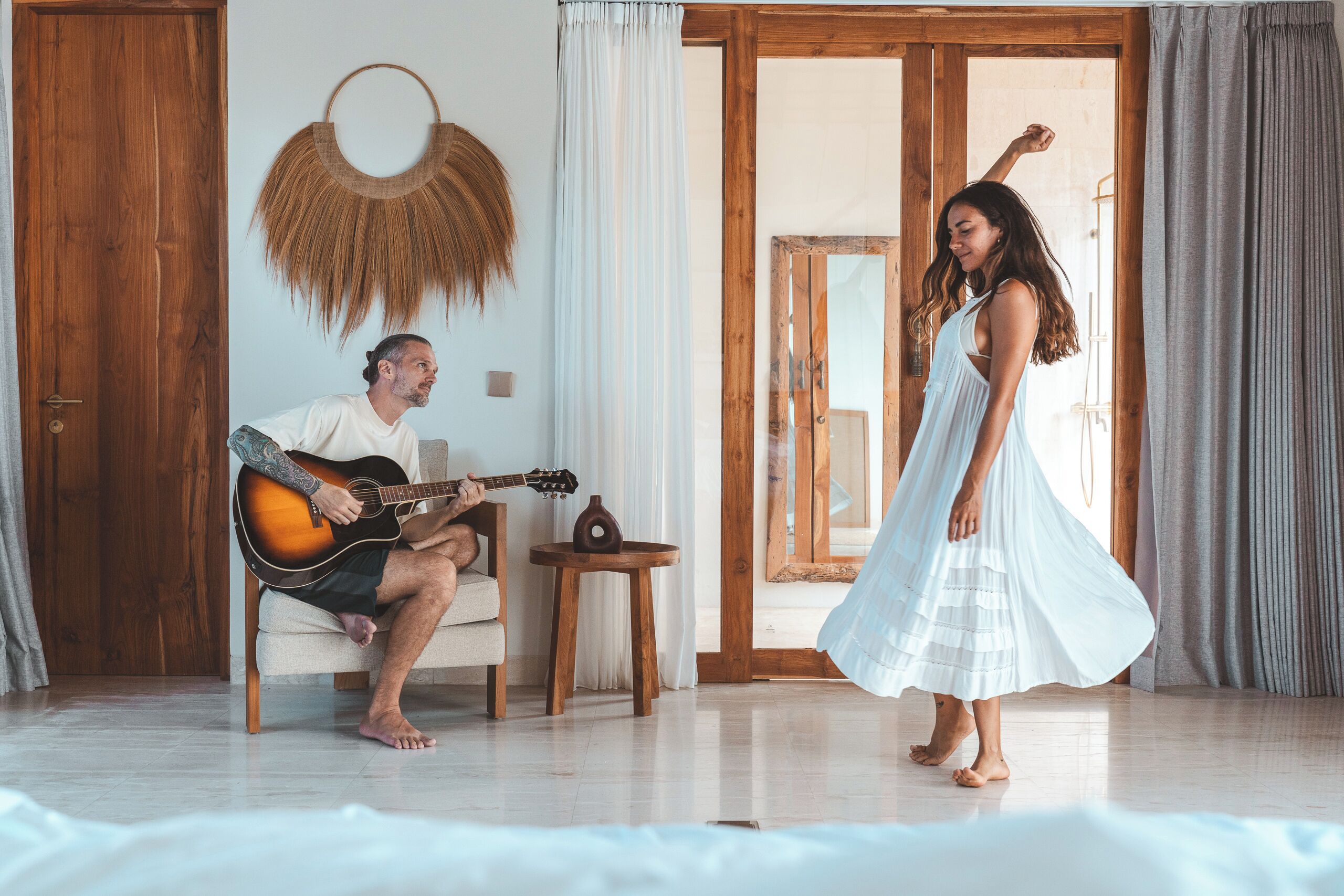 A room with a wooden door, a guitar, a small table with a vase, and a woman in a flowing dress.