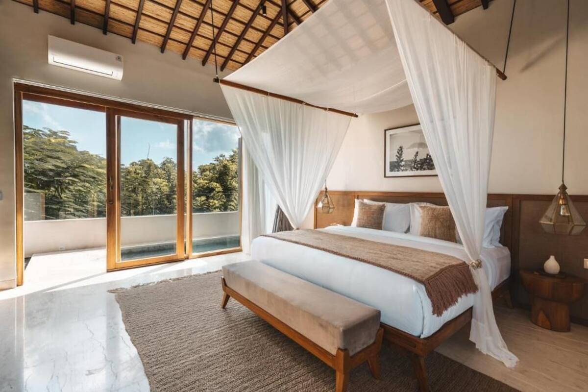 Un dormitorio con cama con dosel, una banca, un ventanal grande y un cuadro en la pared.