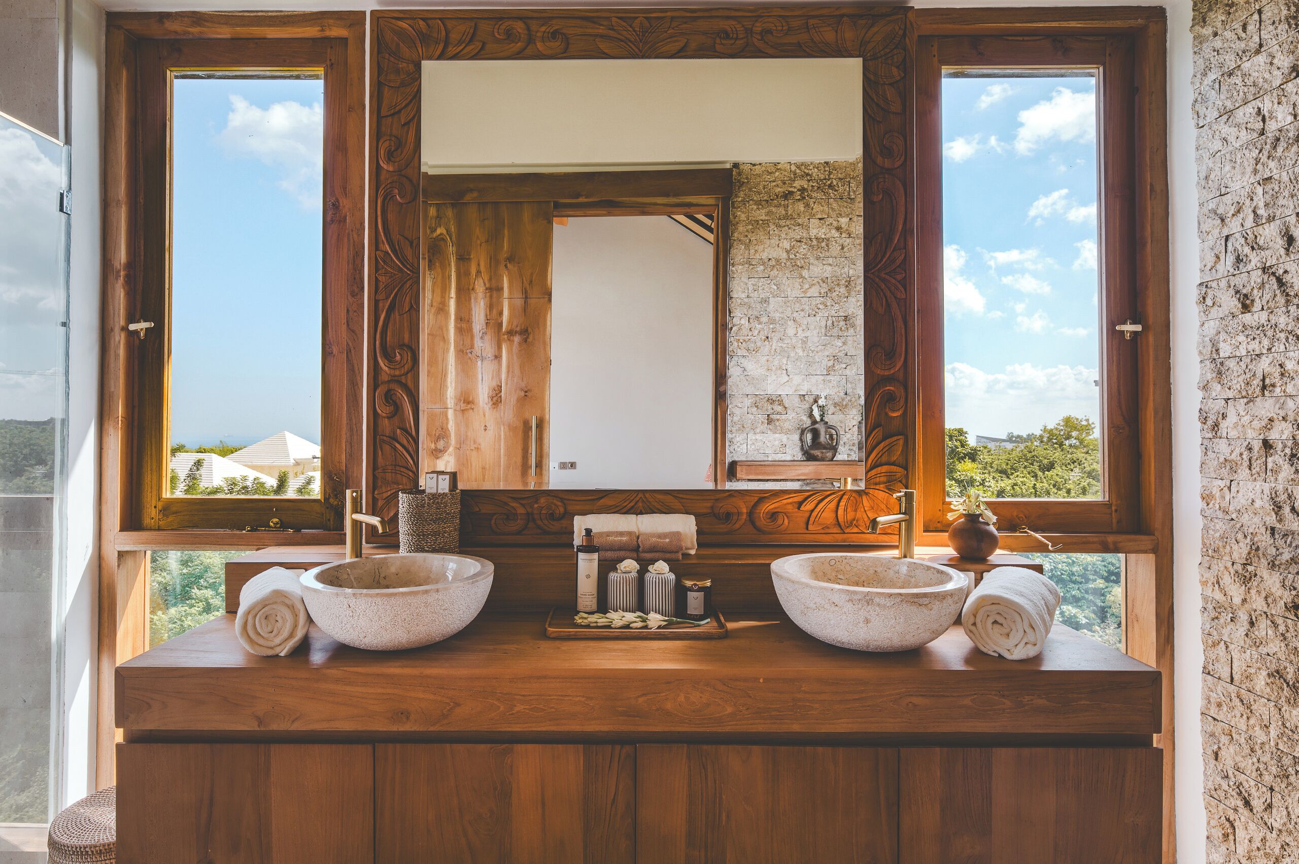 A bathroom with a wooden vanity, two stone sinks, a large mirror, and a view of greenery outside.