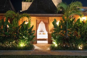 A well-lit entrance to a building with a bed and canopy, surrounded by lush tropical plants.