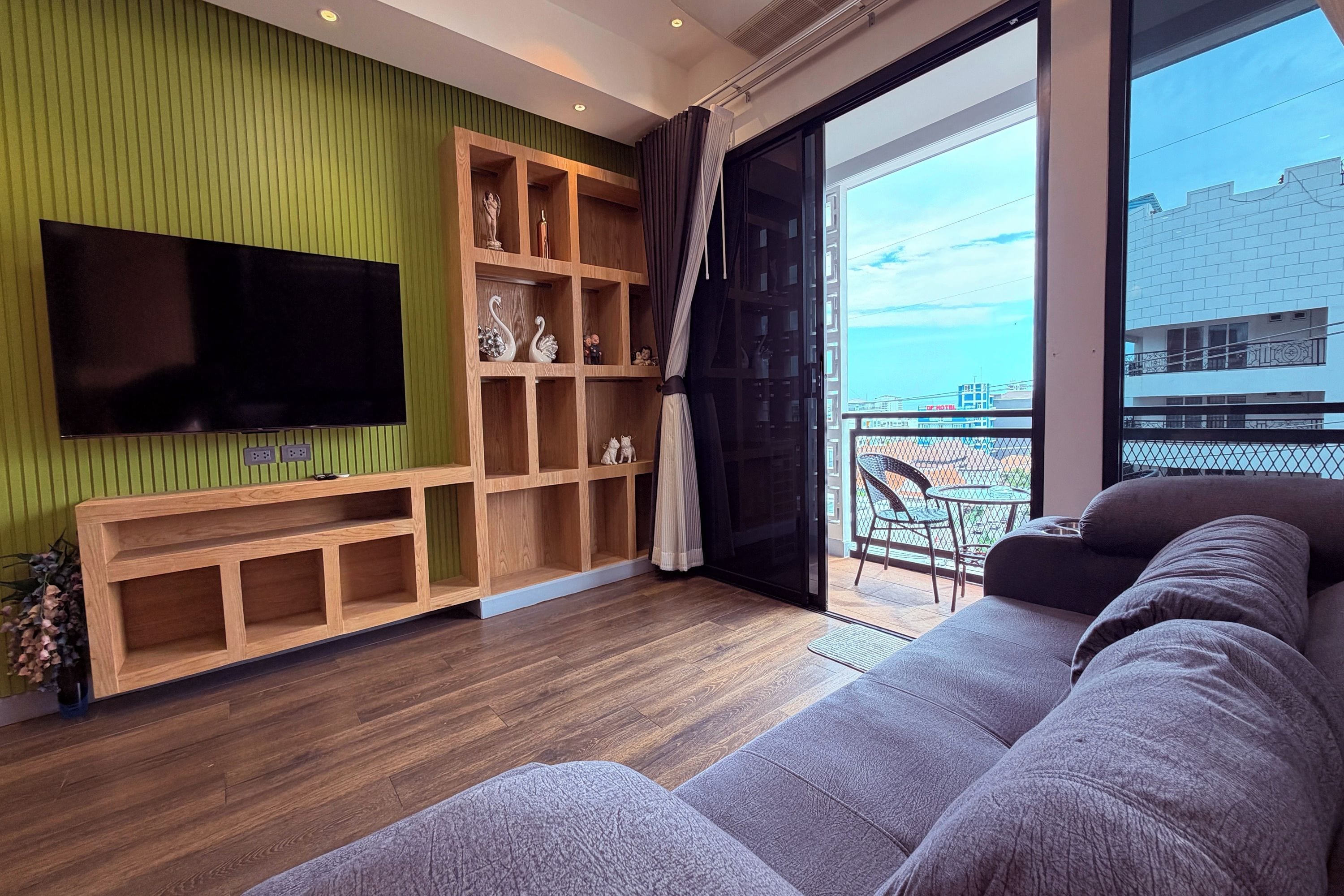 A modern living room with a flat-screen TV, wooden shelving, a grey sofa, and a balcony with a view of the sea.