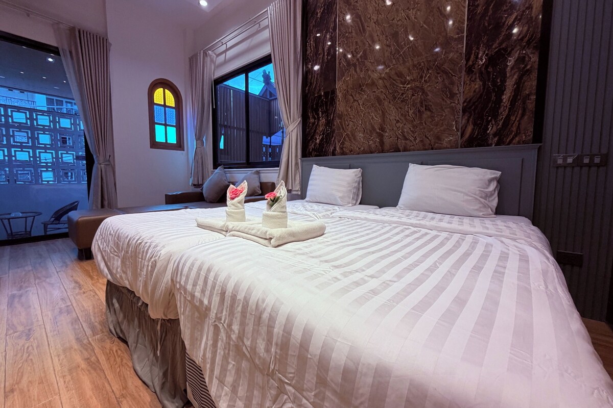 A hotel room with a large bed, a window with curtains, a marble wall, and a view of buildings outside.