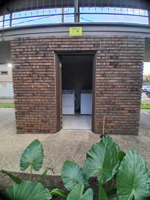 A small brick building with a sign that says 'SMILE! YOU'RE ON CAMERA' above the entrance. Inside, there are two washing machines. - Quiet 2 Bedroom Boutique Style Apt in the Heart of Mid City Baton Rouge  (Baton Rouge)
