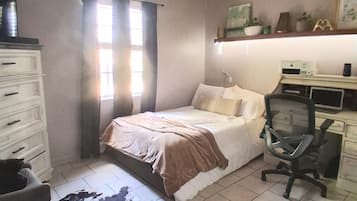 Een slaapkamer met een bed, een bureau, een stoel, een plafondventilator, een raam met gordijnen, en een koeienhuidtapijt.