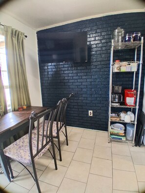 A dining area with a dark brick wall, a wooden table, and chairs. A metal shelf with kitchen items is against the wall. - Quiet 2 Bedroom Boutique Style Apt in the Heart of Mid City Baton Rouge  (Baton Rouge)