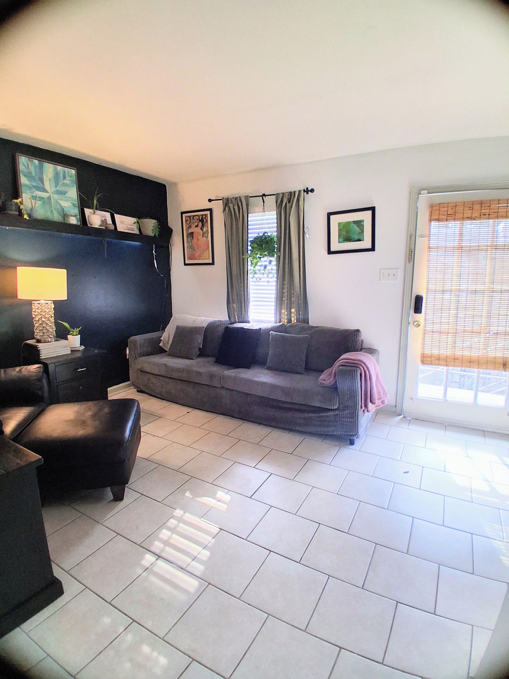 A living room with a sofa, a coffee table, and a lamp. There is a door with a window and a plant on the windowsill.