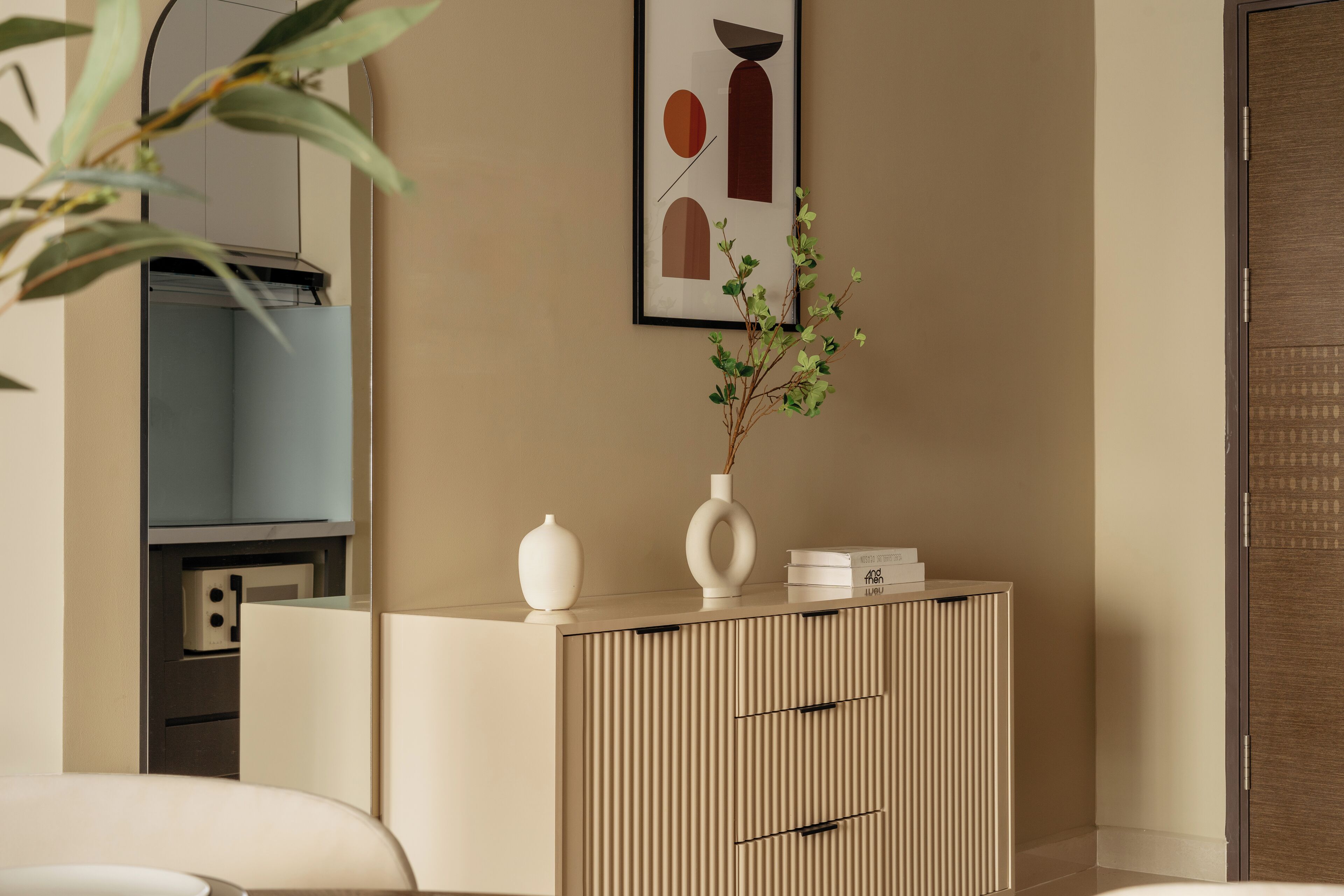A modern interior with a beige wall, a framed abstract artwork, a wooden cabinet with drawers, and a potted plant.