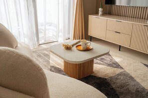 Ein modernes Wohnzimmer mit einer Couch, einem Couchtisch mit Tasse und Pflanze, einem Fernseher auf einer Holzkommode und leichten Vorhängen.