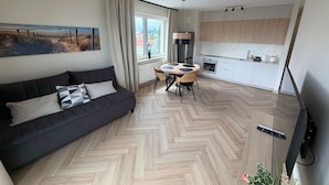 Un salon moderne avec un canapé noir, une table à manger avec des chaises, un coin cuisine avec des meubles blancs et une grande fenêtre avec des rideaux.