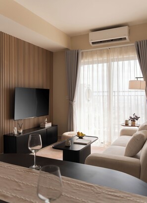Ein modernes Hotelzimmer mit Flachbildfernseher, einem Esstisch mit Obstschale, einer Couch und einem großen Fenster mit leichtem Vorhang.