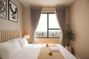 Ein Hotelzimmer mit Bett, einem Fenster mit Vorhängen, Blick auf die Stadt, einer Zimmerpflanze und gerahmten Bildern an der Wand.