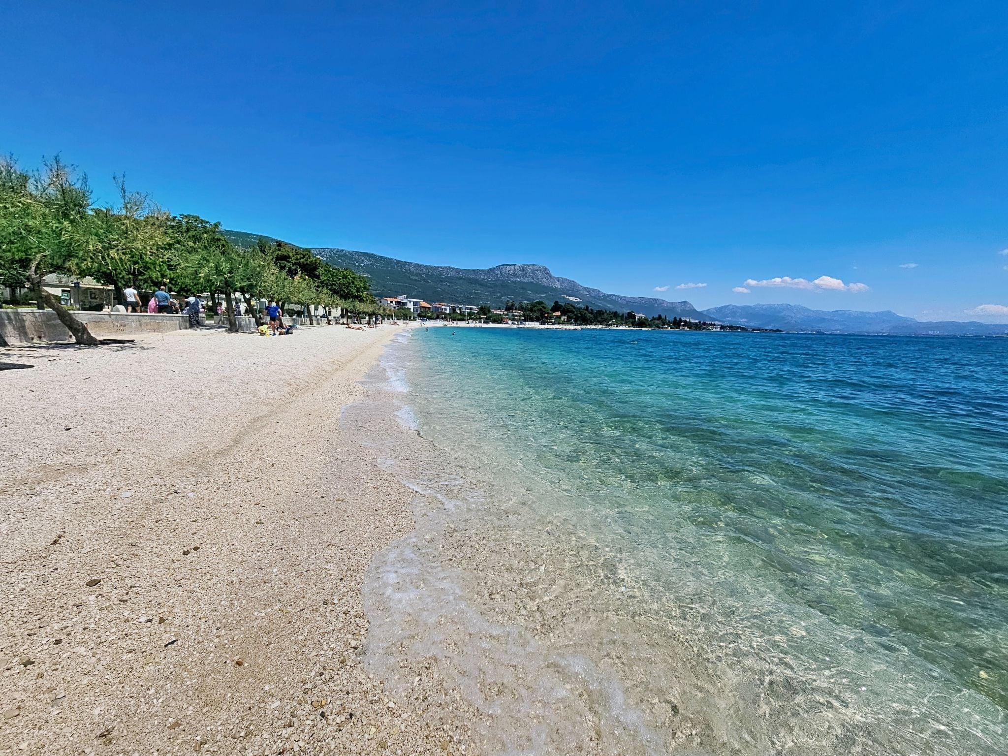 Una playa con aguas cristalinas, una orilla arenosa y edificios al fondo.