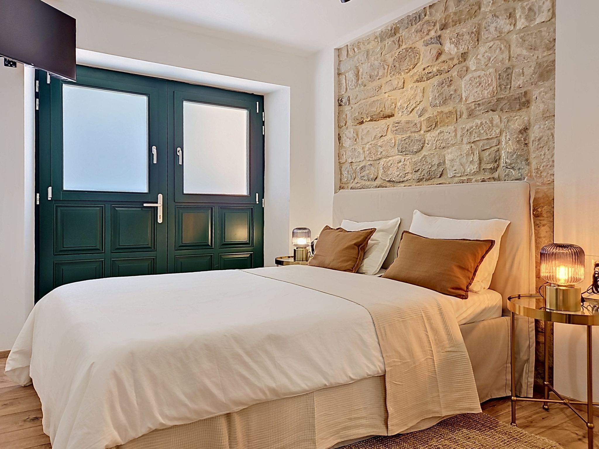A bedroom with a large bed, a stone wall, a door, and a nightstand with a lamp.