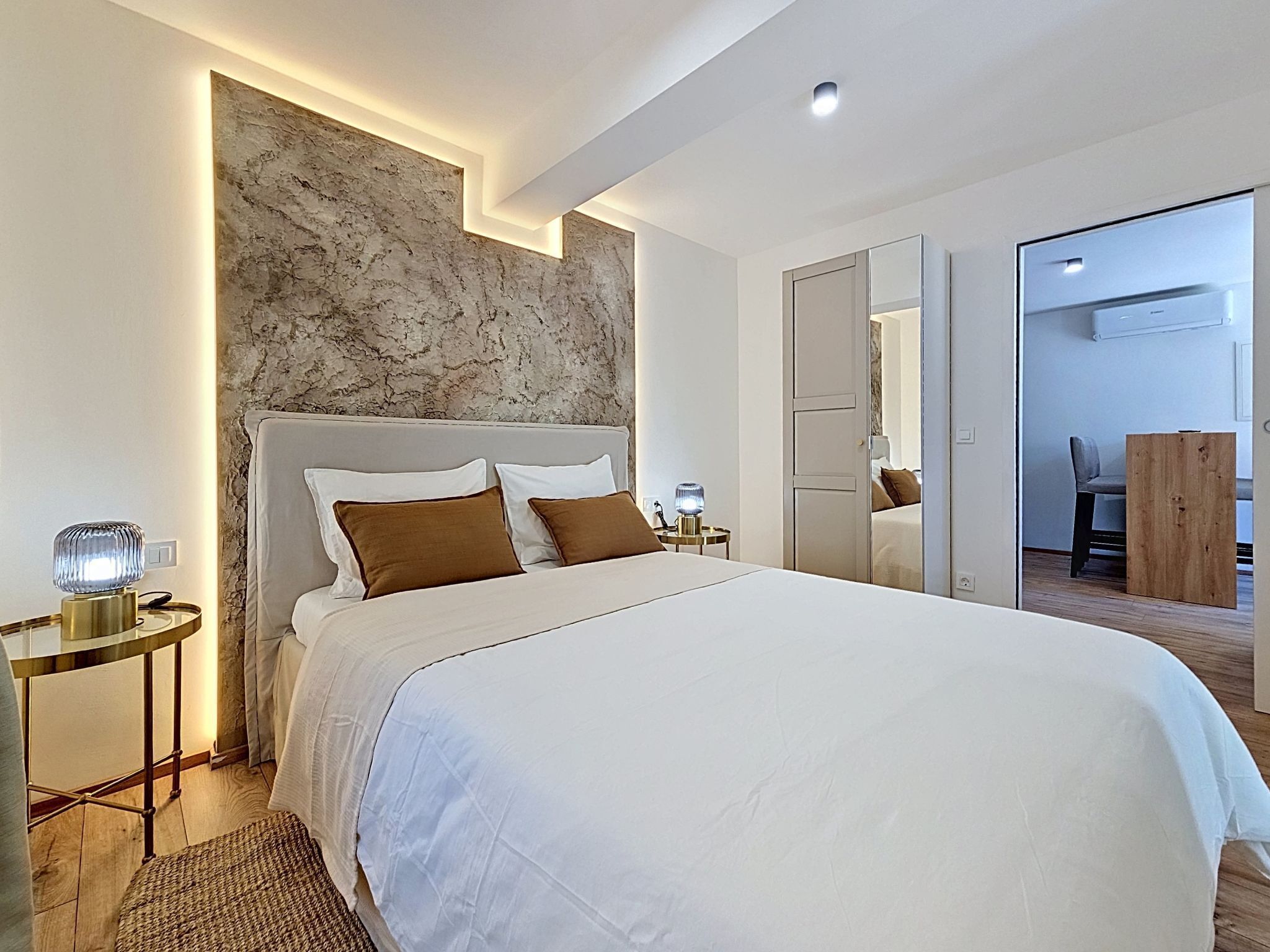 Une chambre moderne avec un grand lit, un mur décoratif en marbre, une table de chevet avec une lampe et une petite table en bois.