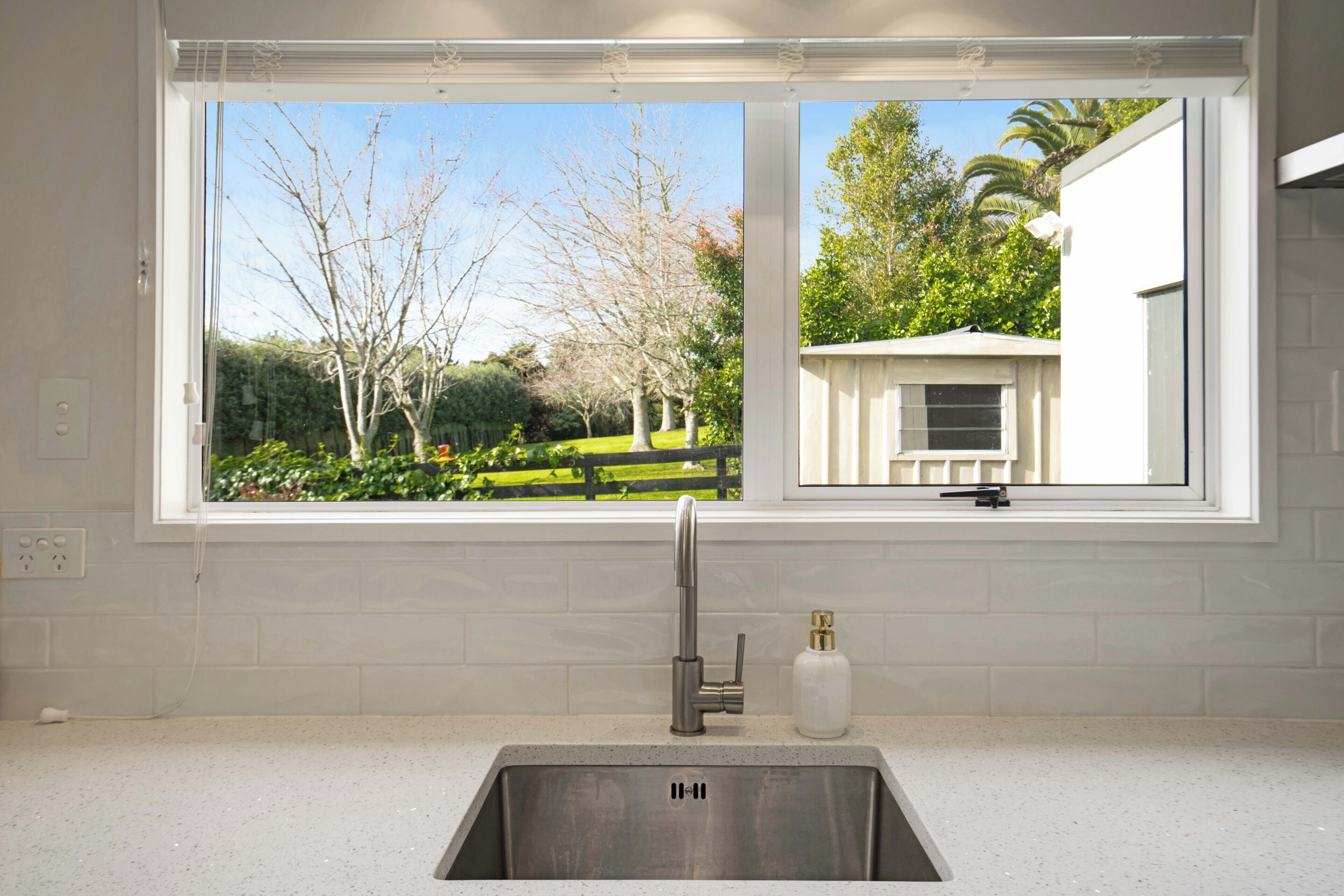 A kitchen with a stainless steel sink, a window with a view of a garden, and a white tiled backsplash.
