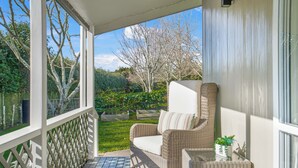 Een overdekt terras met een rieten bank, een klein tafeltje met een plant en uitzicht op een tuin en bomen.