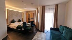 Una habitación de hotel moderna con una cama, dos sillones, un sofá, un armario de madera y un ventanal con cortinas.