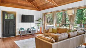 Een moderne woonkamer met een houten plafond, een grote bank, een televisie en een raam met uitzicht op bomen.