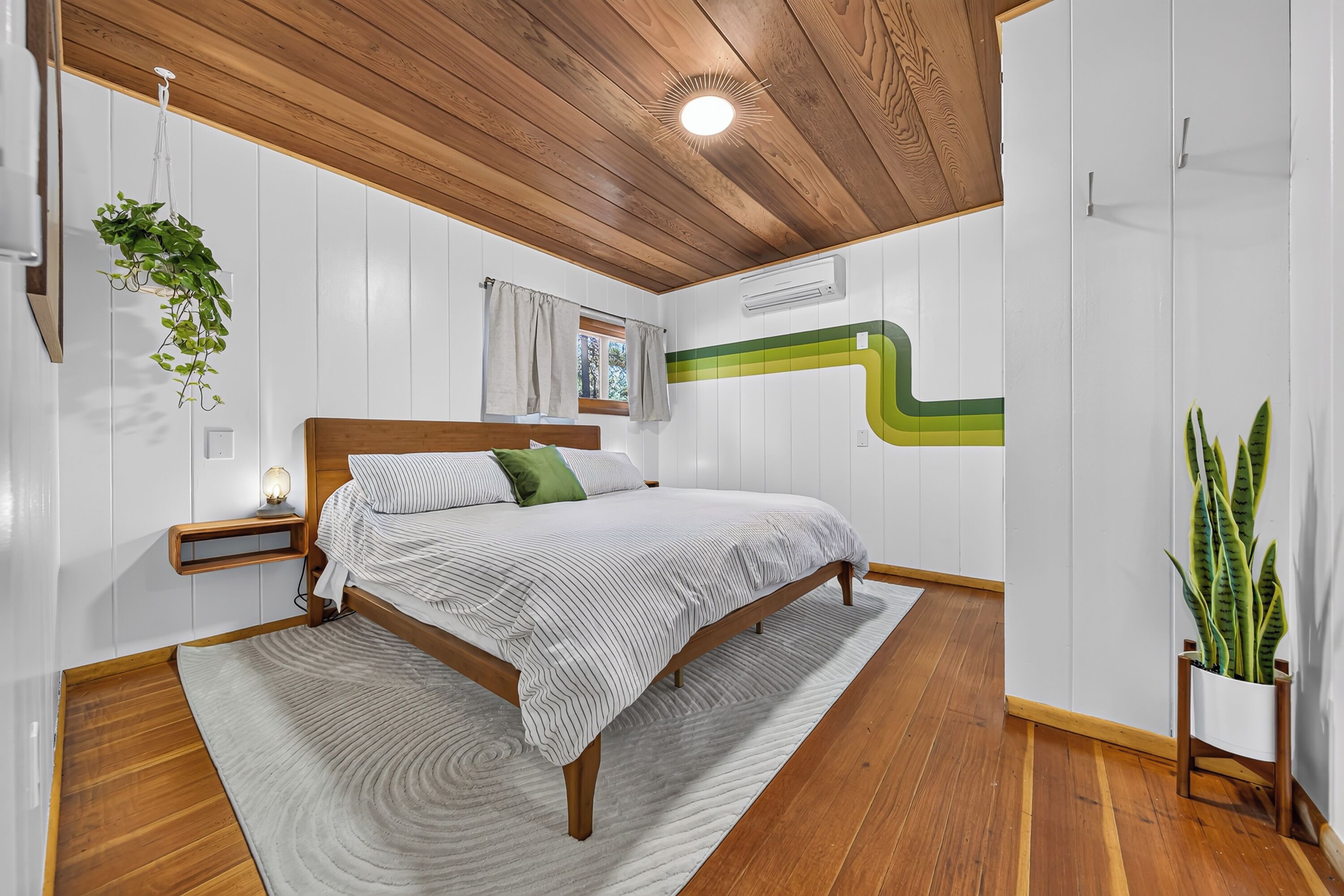 A bedroom with a wooden ceiling, a bed with a striped bedspread, a bedside table with a lamp, and a green plant.