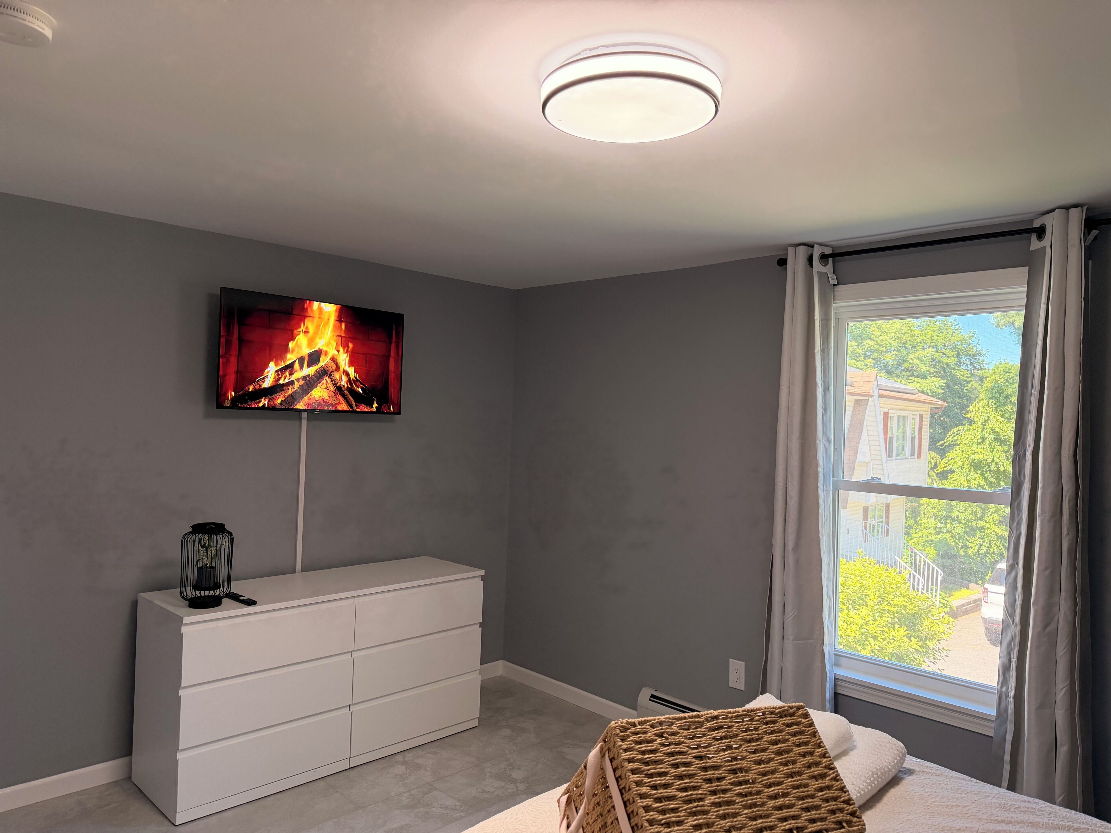 A bedroom with a TV on the wall, a white dresser, a window with curtains, and a view of greenery outside.