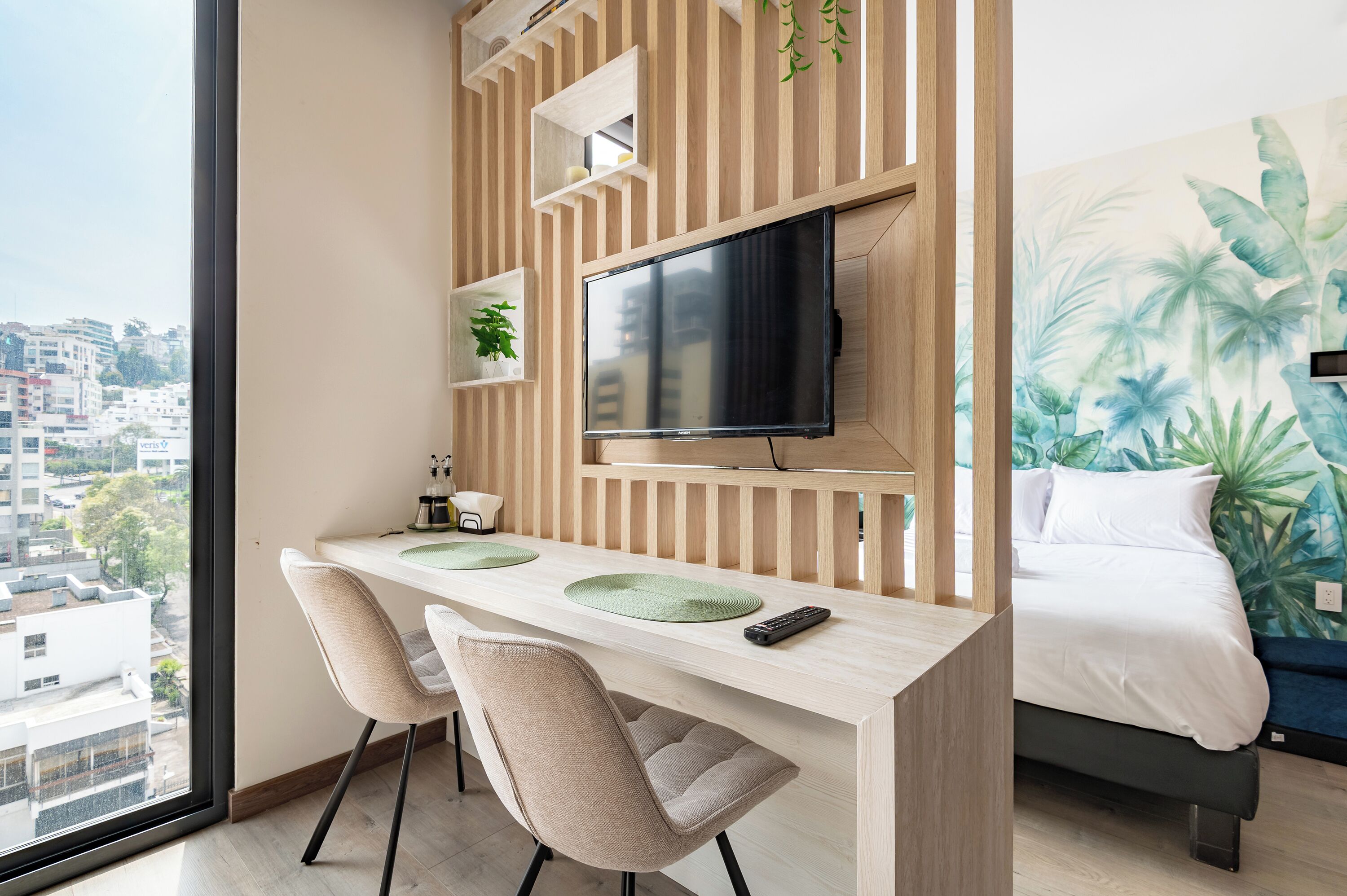 Een moderne hotelkamer met een groot raam, een houten bureau met een flatscreen televisie, twee stoelen, een bed en een muurschildering met een tropisch thema.