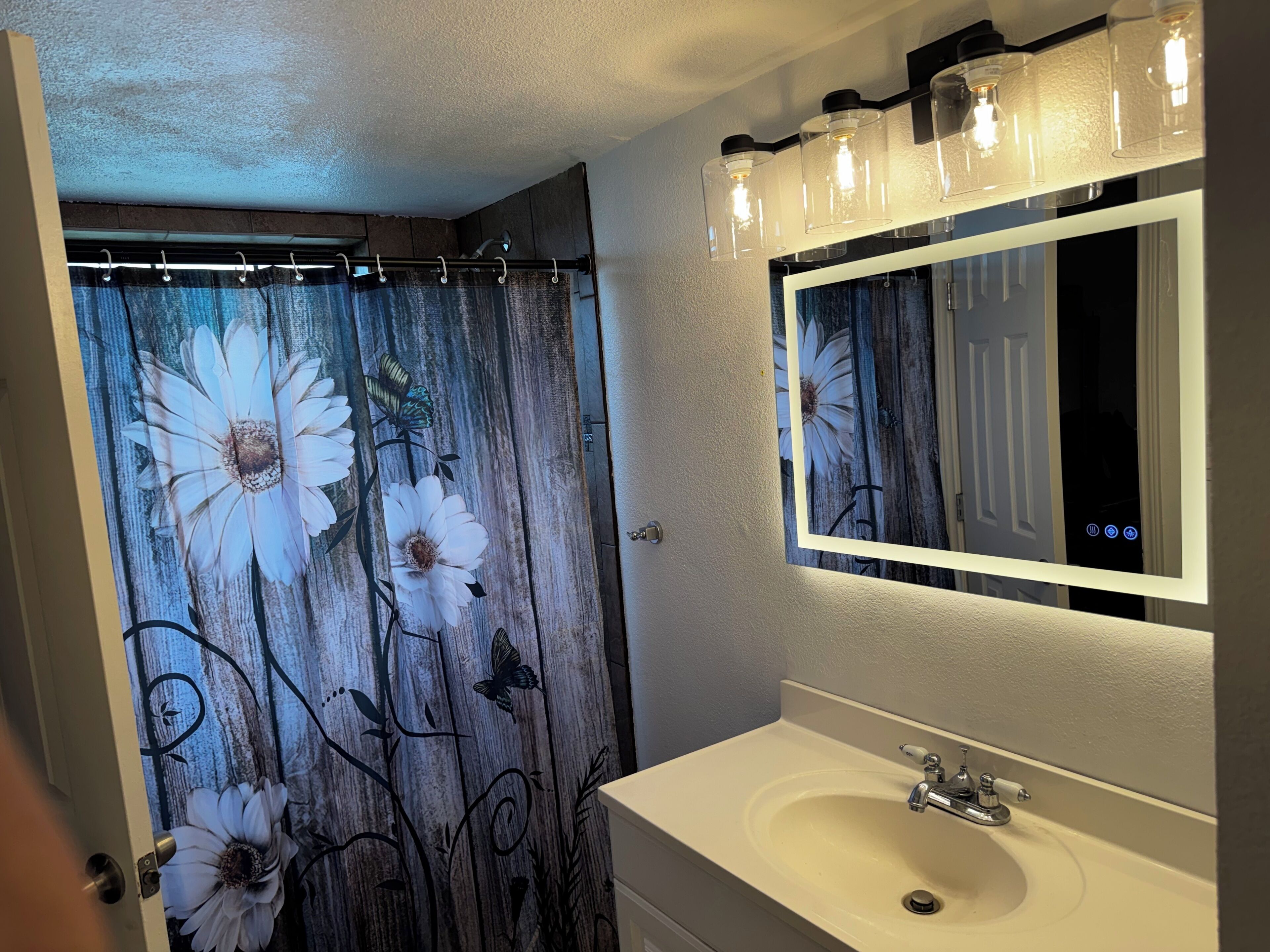 Un baño con cortina de ducha decorada con motivos florales y mariposas, un lavamanos blanco con un espejo encima y una lámpara con tres bombillas.