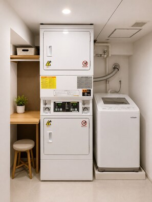A compact laundry room with a stacked washer and dryer, a small stool, and a shelf with a plant. - BACOWA AWAZA (Nishi-ku, Osaka)