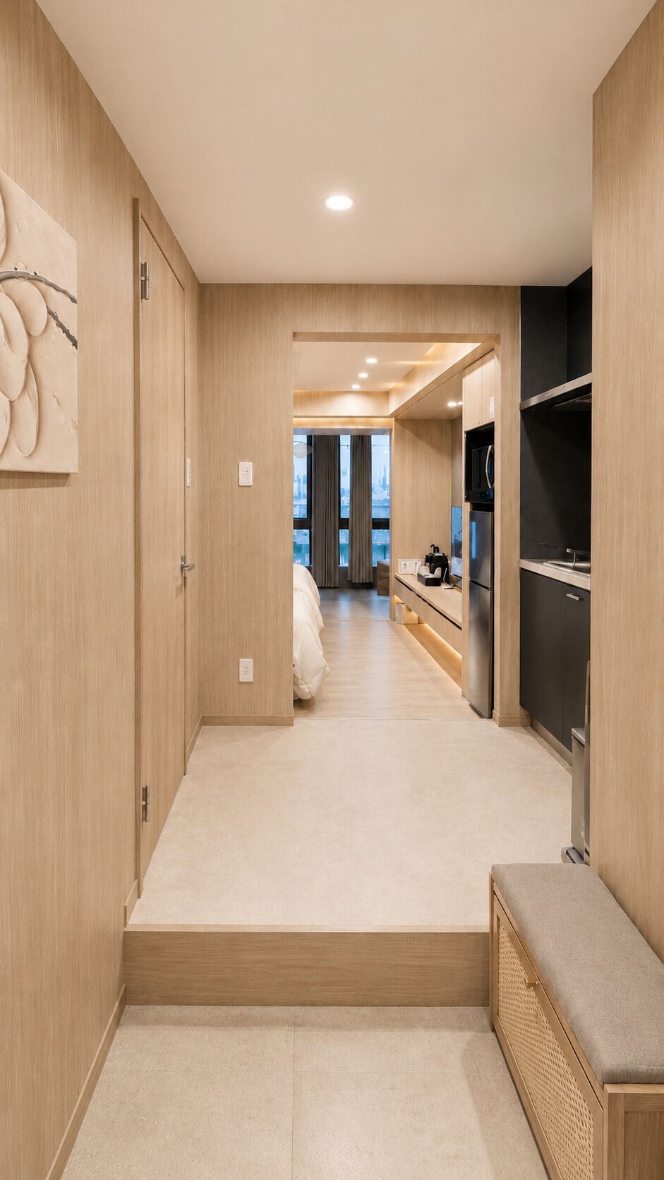 A modern hotel room with a wooden floor, a bed, a kitchenette, and a view of the cityscape.