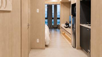 A modern hotel room with a wooden floor, a bed, a kitchenette, and a view of the cityscape.