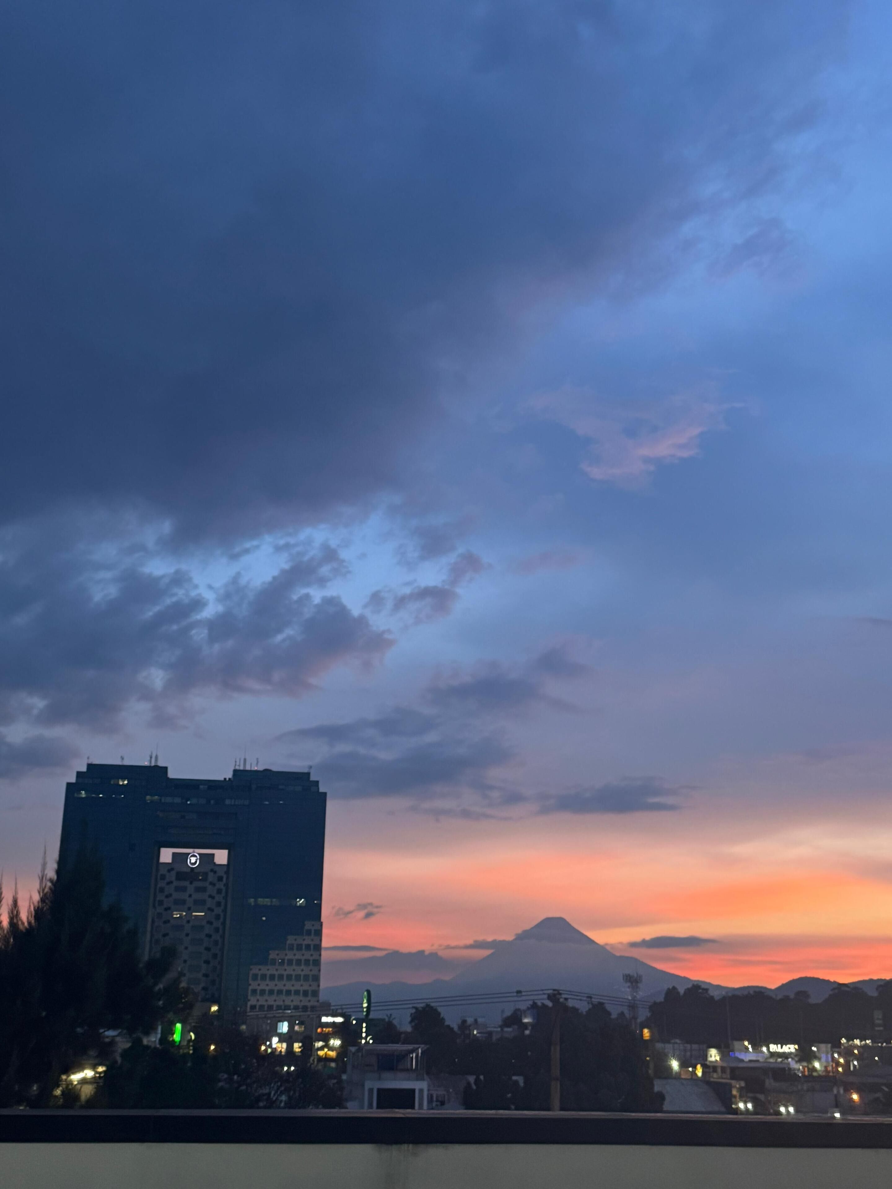 Un paisaje urbano al anochecer con un edificio destacado que tiene un ventanal circular y una montaña al fondo.