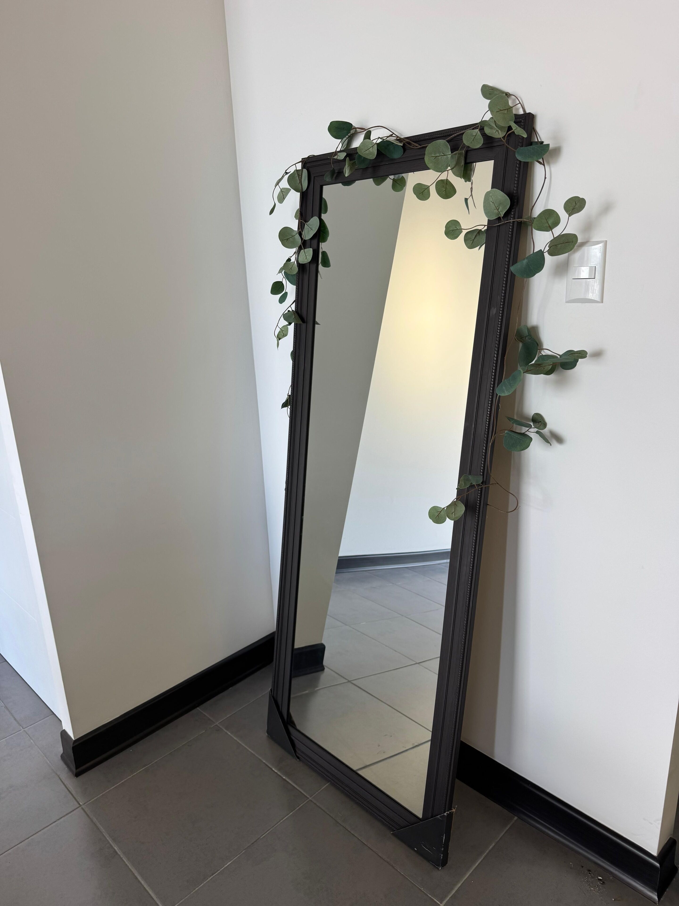 Um espelho alto com moldura escura e detalhes de folhagem verde, pendurado em uma parede branca.