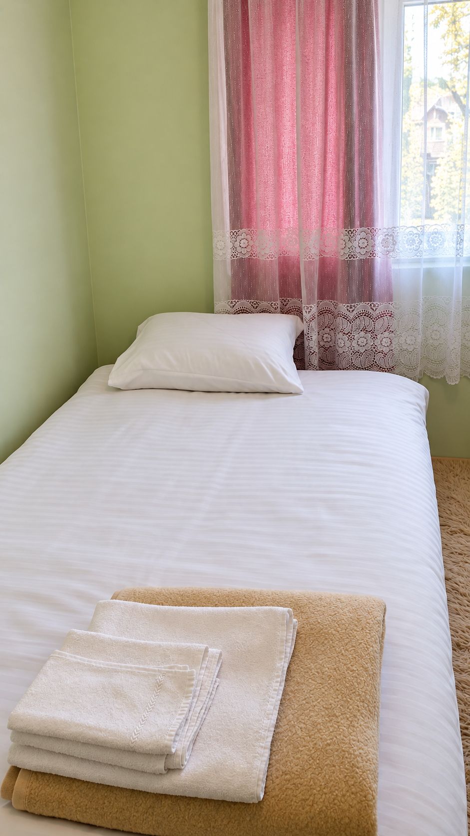 Una cama individual con ropa de cama blanca, una alfombra color arena, una almohada blanca y una toalla blanca doblada.